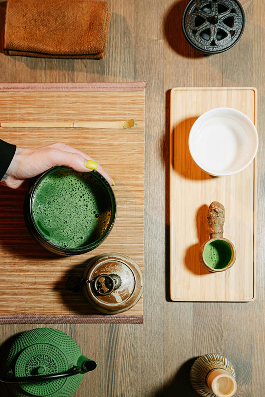 Eine Person hält eine schwarze Schale mit aufgeschäumtem, grünen Matcha, umgeben von einer Bambusmatte, einer weißen Schale, einem kleinen Gefäß mit Matcha sowie verschiedenen Tee-Utensilien auf einem Holztisch in Vogelperspektive