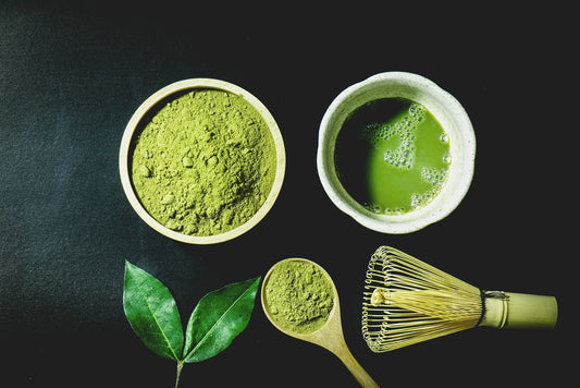 Matcha-Pulver in Holzschale und Löffel neben aufgebrühtem Tee in Keramikschale mit Bambusbesen (Chasen) und Teeblatt