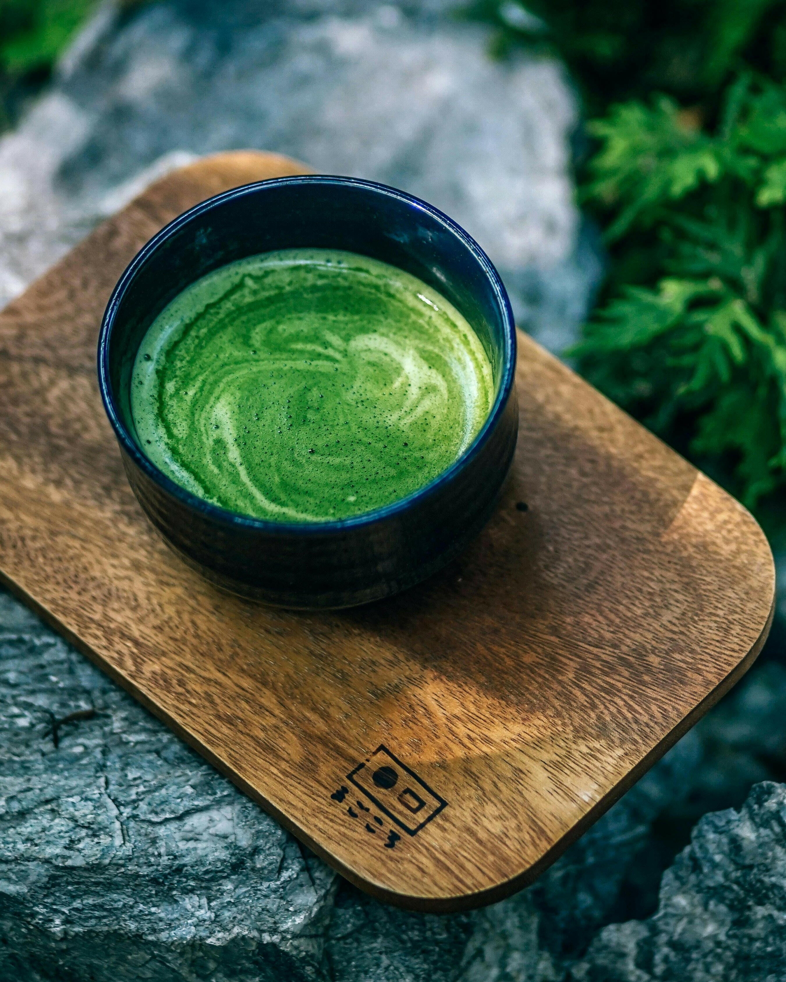 Eine dunkle Schale mit schaumigem, grünen Matcha steht auf einem hölzernen Tablett, das auf grauen Steinen ruht und von üppigem Grün umgeben ist.
